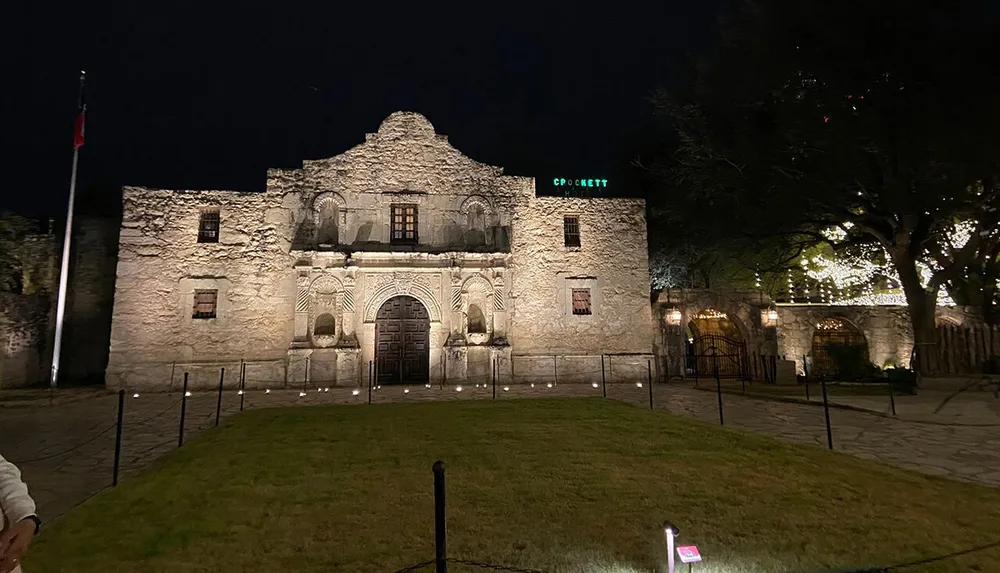 The image shows the Alamo Mission illuminated at night with a large Texas flag flying on the left and a neon sign above the right side of the complex