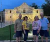 Four individuals are posing for a photo in the evening in front of the historic Alamo mission building