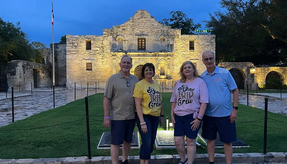 Four individuals are posing for a photo in the evening in front of the historic Alamo mission building