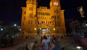 The image shows a group of people walking towards a grand, illuminated building at night, with a blurred effect suggesting motion or a low-light capture.