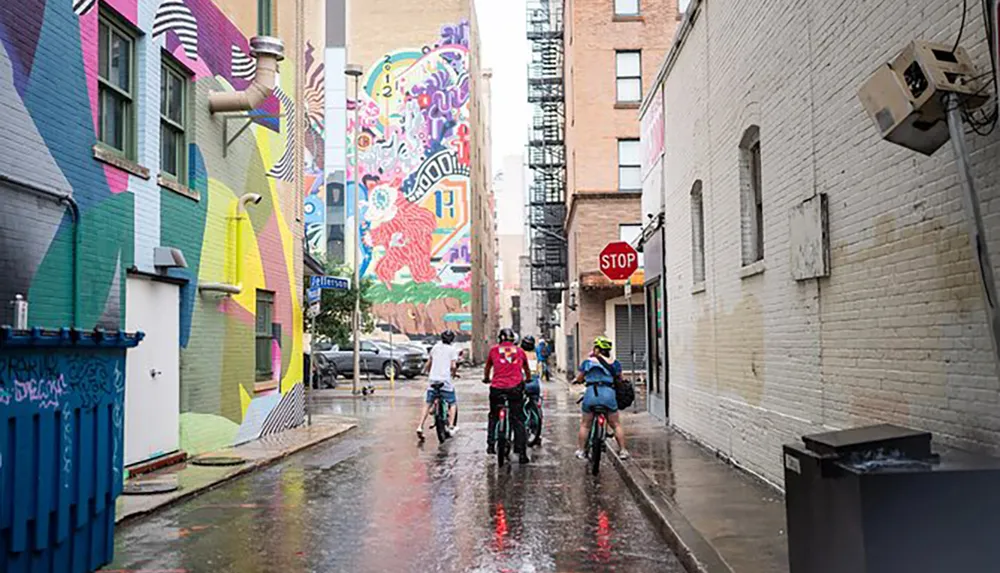 Cyclists ride through an urban alley adorned with vibrant street art after a rain