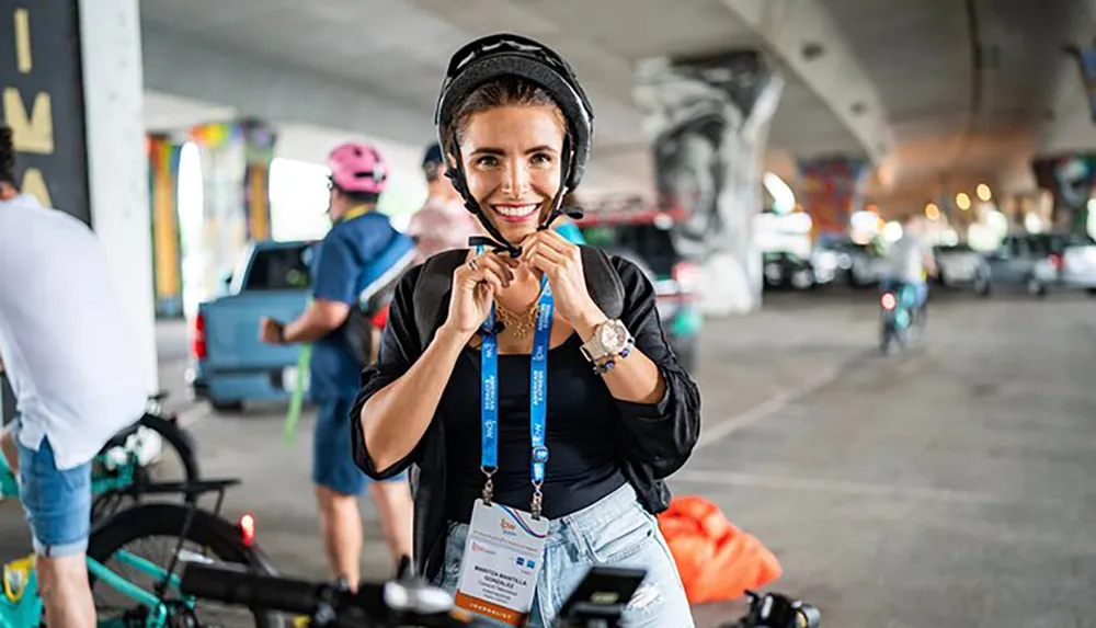 A woman with a cheerful expression is fastening her bicycle helmet ready to ride amidst other cyclists in an urban setting under a bridge