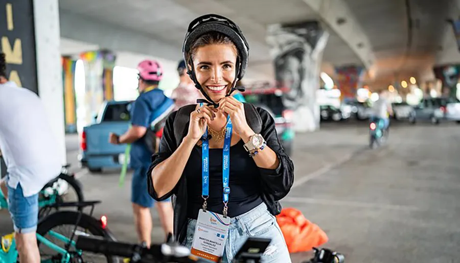 A woman with a cheerful expression is fastening her bicycle helmet, ready to ride, amidst other cyclists in an urban setting under a bridge.