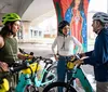 Three cyclists wearing helmets pause for a conversation under an overpass with colorful murals in the background