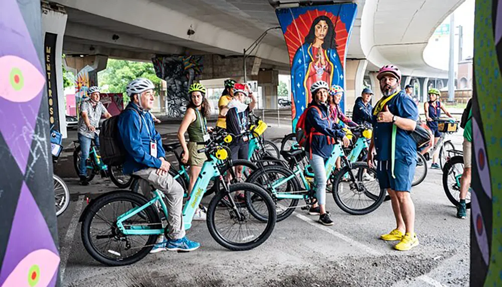A group of cyclists wearing helmets is gathered under an overpass adorned with colorful street art