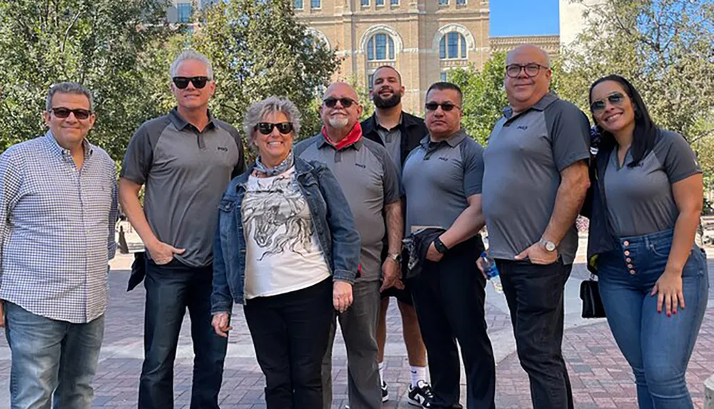 A group of eight smiling people poses for a photo outdoors on a sunny day with trees and a large building in the background