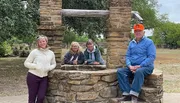 Four people are posing for a photo at an old stone well, with three grouped together inside the well's frame and one standing to the side.