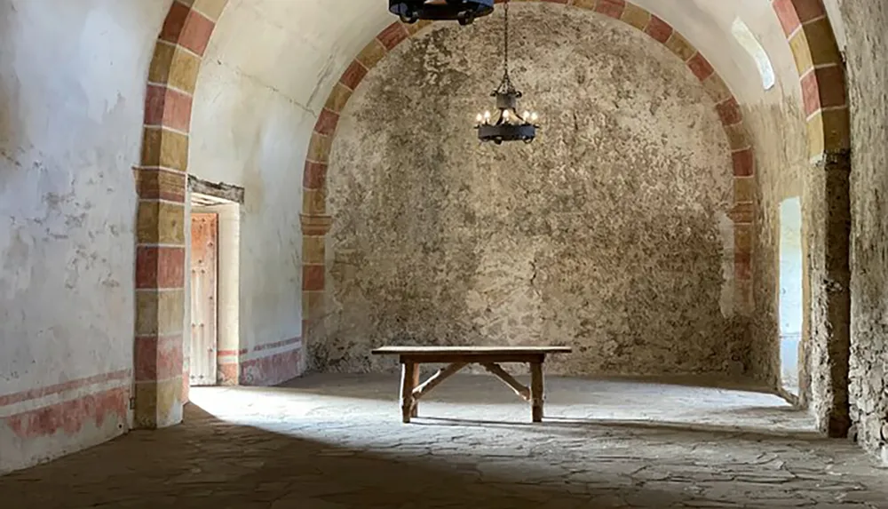 An old arched room with a solitary wooden table fading wall decorations and a hanging metal chandelier evoking a sense of historical austerity