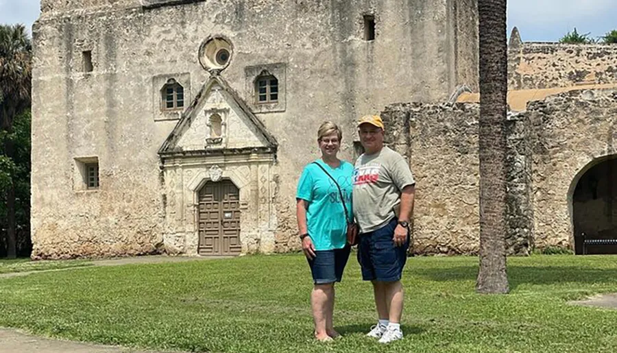 A couple is posing for a photo in front of an old, historic stone building with a detailed façade under a clear sky.