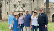 A group of six individuals are posing for a photo in front of an old stone building with historical architecture.