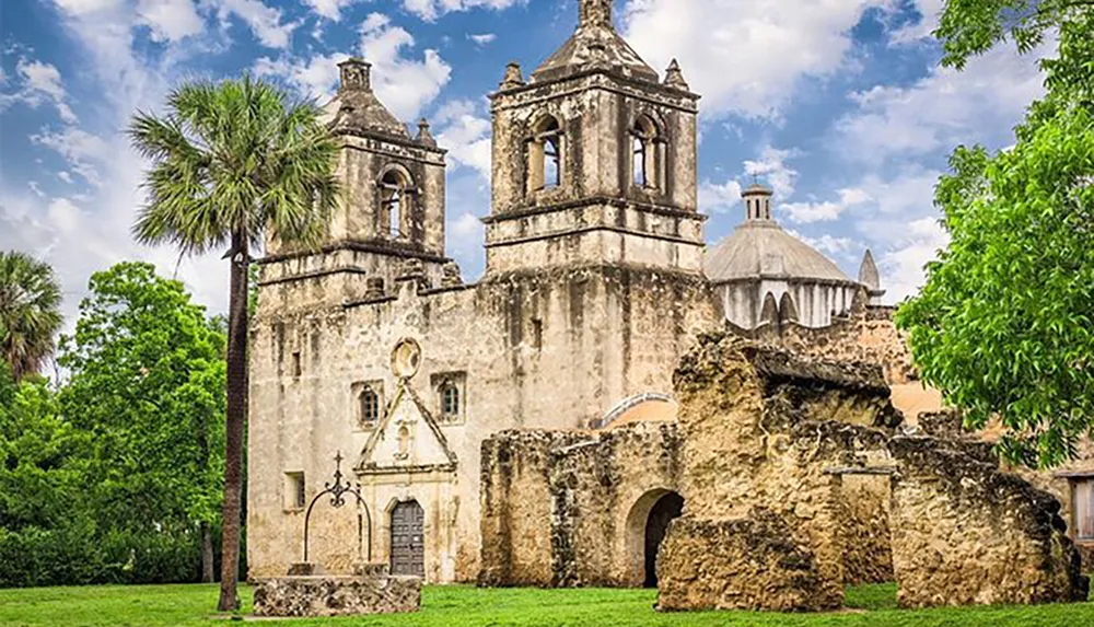 The image depicts the weathered facade of an old stone mission with twin bell towers surrounded by lush greenery under a partly cloudy sky