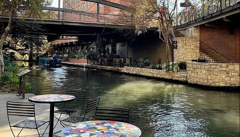 The image showcases a scenic view of a riverwalk with a boat carrying passengers an outdoor dining area with mosaic-topped tables and a pedestrian bridge overhead all of which create a tranquil urban outdoor setting