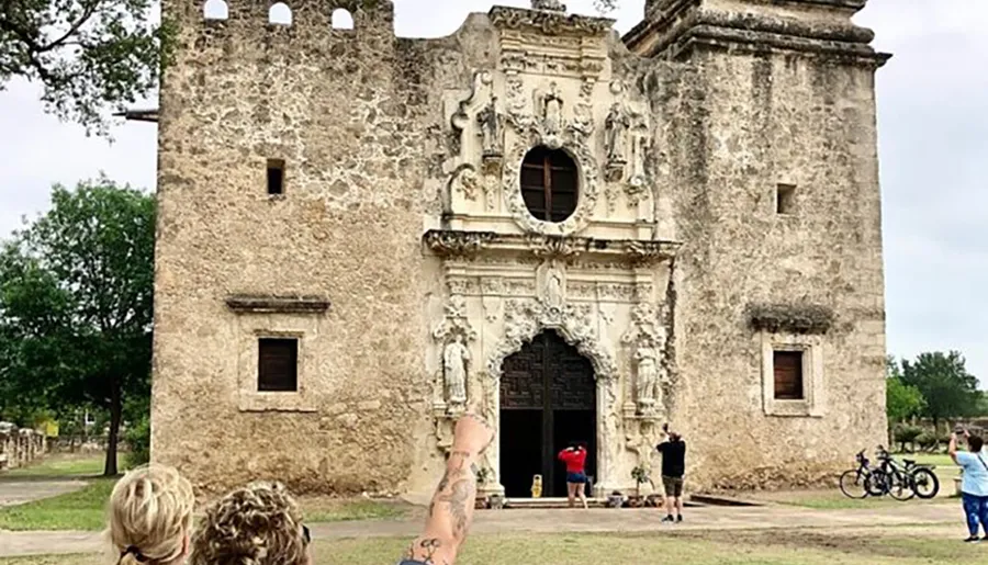 Visitors are exploring and capturing photos of a historic stone facade with ornate carvings, possibly of a mission or church, under a cloudy sky.