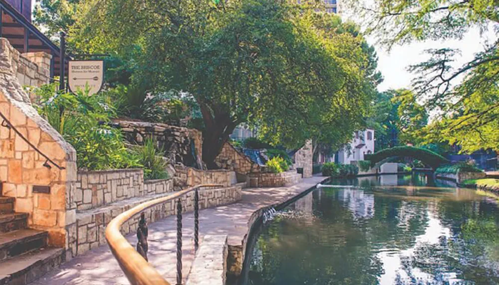 This image depicts a tranquil river walk with lush greenery stone paths railings and a sign for The Briscoe in a serene park-like urban setting