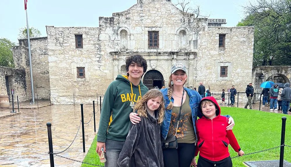 A group of four people are posing for a photo in front of the Alamo on a cloudy day