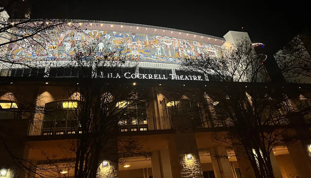 The image shows the illuminated exterior of the Lila Cockrell Theatre at night featuring a large colorful mural above the entrance sign