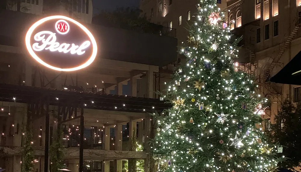 The image shows a large illuminated Christmas tree adorned with lights and ornaments with a neon sign reading Pearl in the background at dusk or night