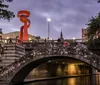 An old stone bridge spans a river in an urban setting illuminated by the glow of streetlights and an orange sculpture at dusk