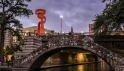 An old stone bridge spans a river in an urban setting, illuminated by the glow of streetlights and an orange sculpture at dusk.