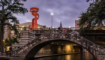 An old stone bridge spans a river in an urban setting, illuminated by the glow of streetlights and an orange sculpture at dusk.