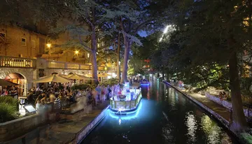 The image shows a vibrant night scene of an illuminated riverwalk with people dining alfresco and a boat gliding on the water.