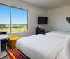 The image shows a modern hotel room with twin beds bright accent pieces like a POW pillow and striped rug a small table and seat by the window and a large window offering a view of the landscape outside
