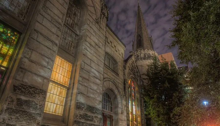 The image shows a historic stone church with stained glass windows illuminated at night under a cloudy sky.