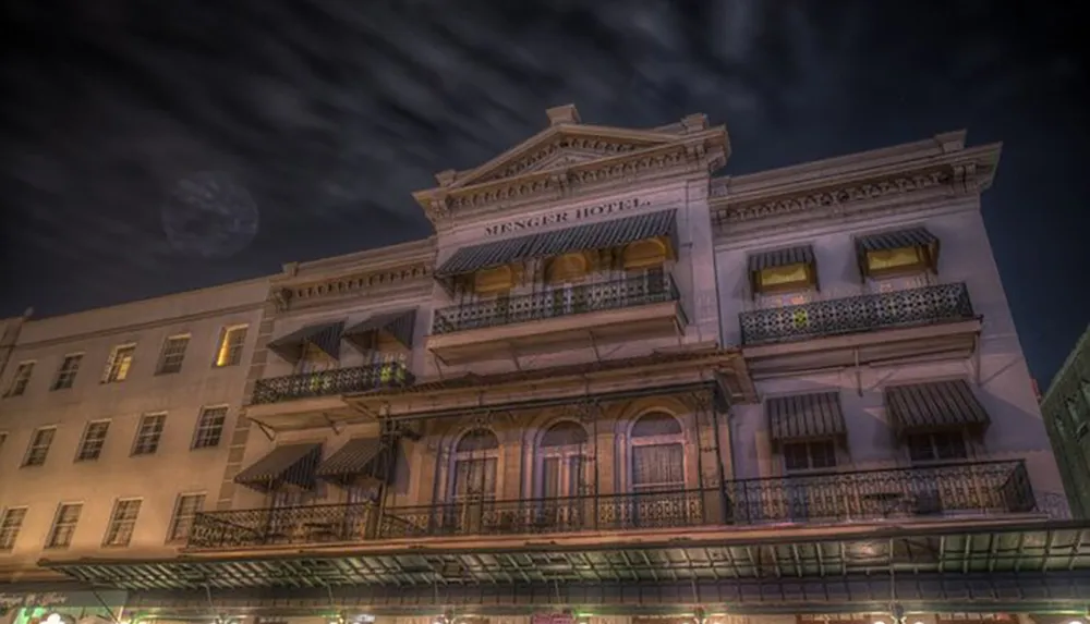 The image shows the historic Menger Hotel at night with a dramatic sky and the moon partially visible through passing clouds