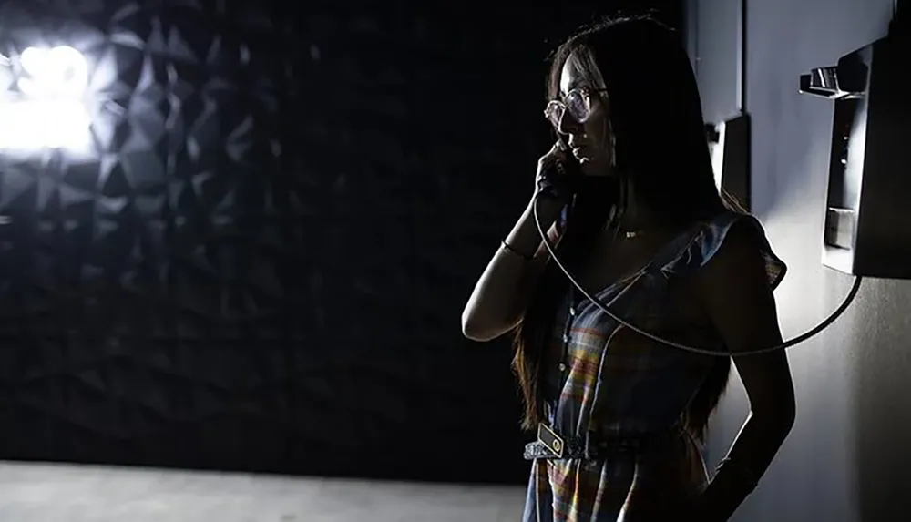 A woman is pictured in a dimly lit room speaking on a payphone while illuminated by a strong light source behind her creating a shadow pattern on the wall