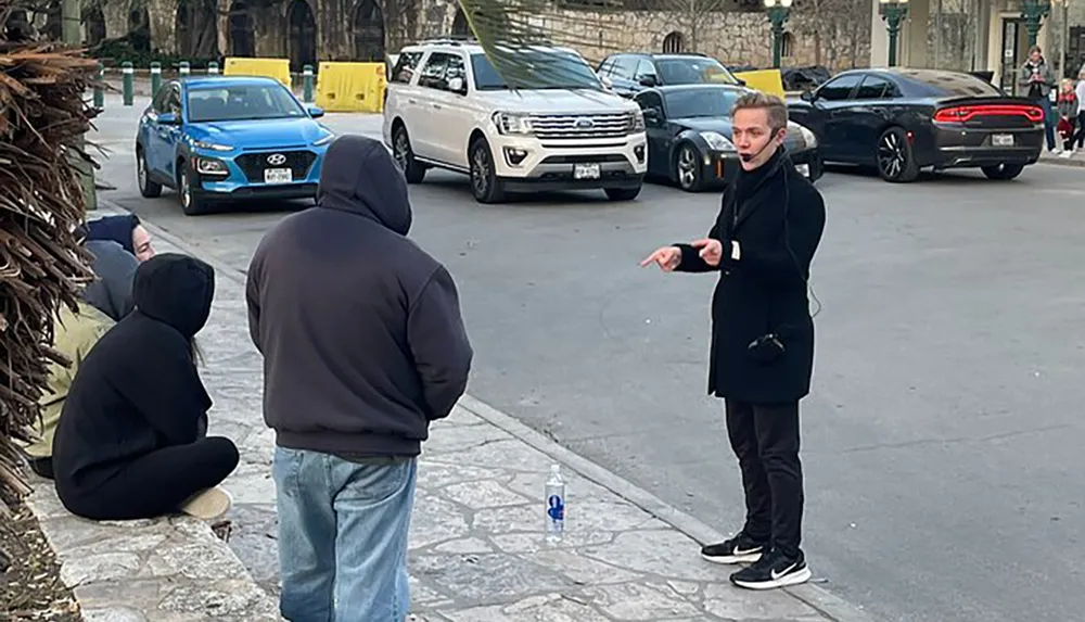 A person appears to be performing an outdoor demonstration or speaking to a small group on the street with a plastic bottle standing upright on the pavement
