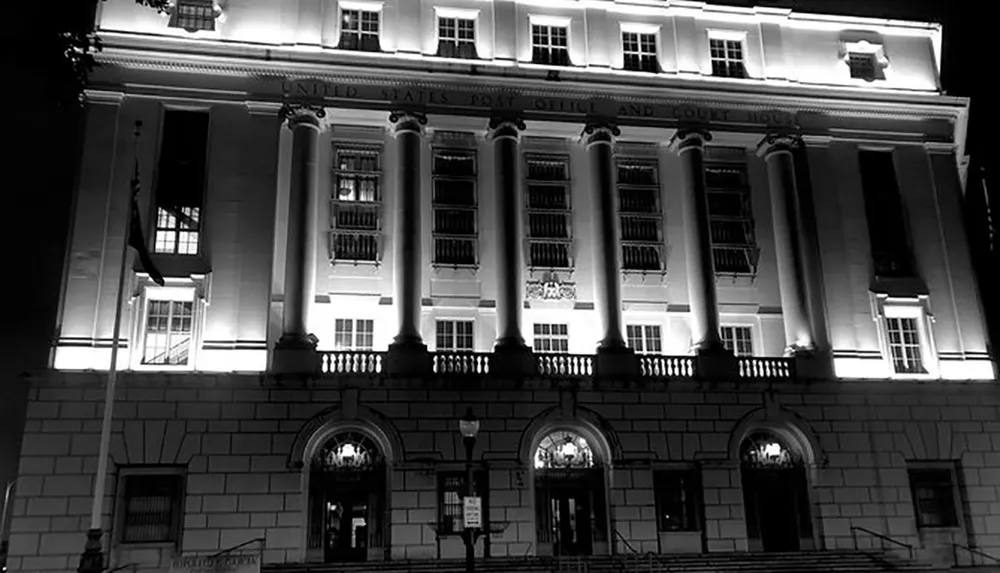 This black and white image depicts the illuminated faade of a grand building at night marked as the United States Post Office and Courthouse