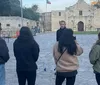 A person is explaining something to a small group of listeners in front of a historic building with a flagpole flying the Texas flag