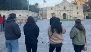 A person is explaining something to a small group of listeners in front of a historic building with a flagpole flying the Texas flag.