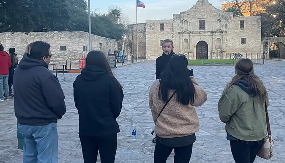 A person is explaining something to a small group of listeners in front of a historic building with a flagpole flying the Texas flag