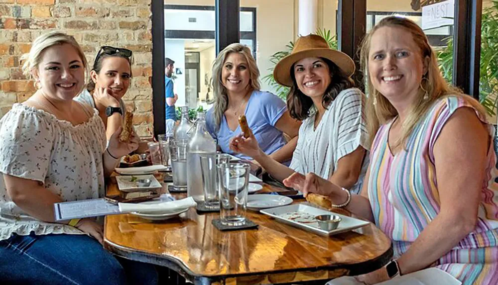 Five women are smiling and posing for a photo while sitting at a wooden table in a restaurant with drinks and small plates of food in front of them