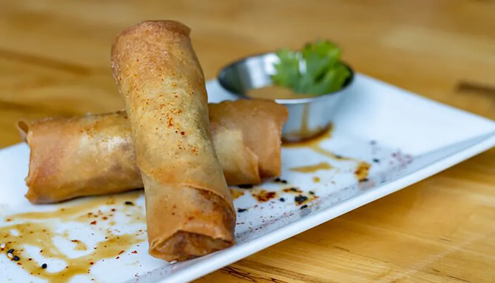 A plate of golden-brown spring rolls is served with a dipping sauce and a garnish of greens on the side
