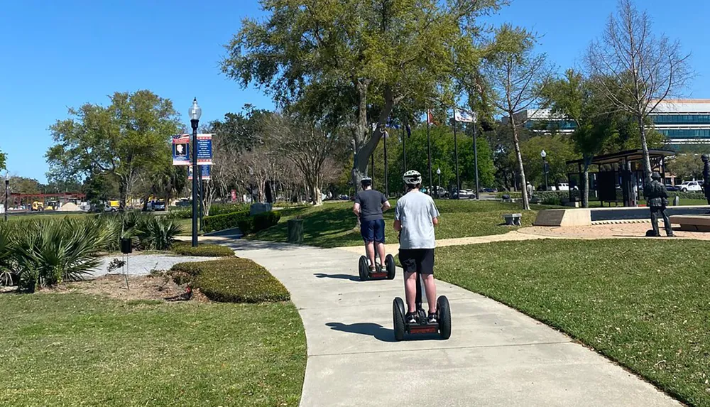 Two individuals are riding Segways along a park path on a sunny day