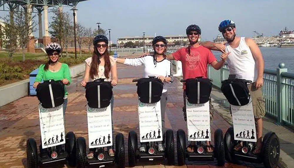 A group of five people are standing on a waterfront boardwalk smiling and posing with their Segways