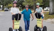 Three people are posing with Segways on a residential street, wearing helmets and smiling, with the signage indicating they are part of a tour group.