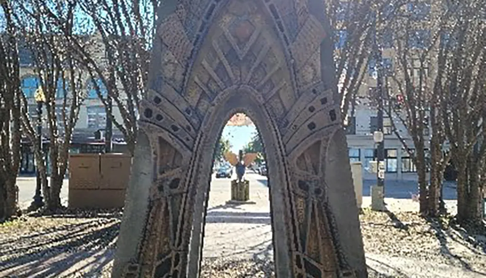 The image shows an intricately designed archway framing a view down a tree-lined pathway with a statue and urban features in the background