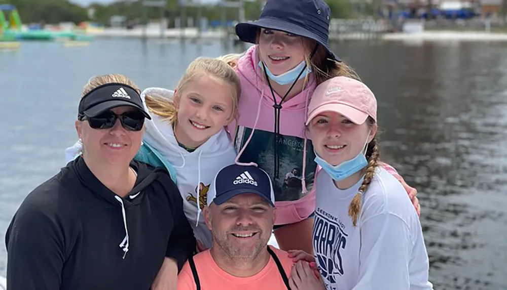 A group of five people likely a family are smiling for a photo by the water with some wearing hats and masks dangling from their ears