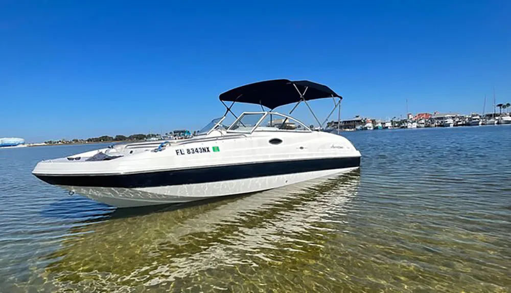 A white motorboat with a canopy is anchored in shallow clear water near the shore with buildings and other boats visible in the distance