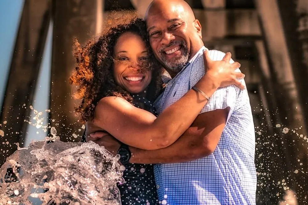 A joyful couple is embracing and smiling in front of a splashing water fountain with sunlight illuminating the scene