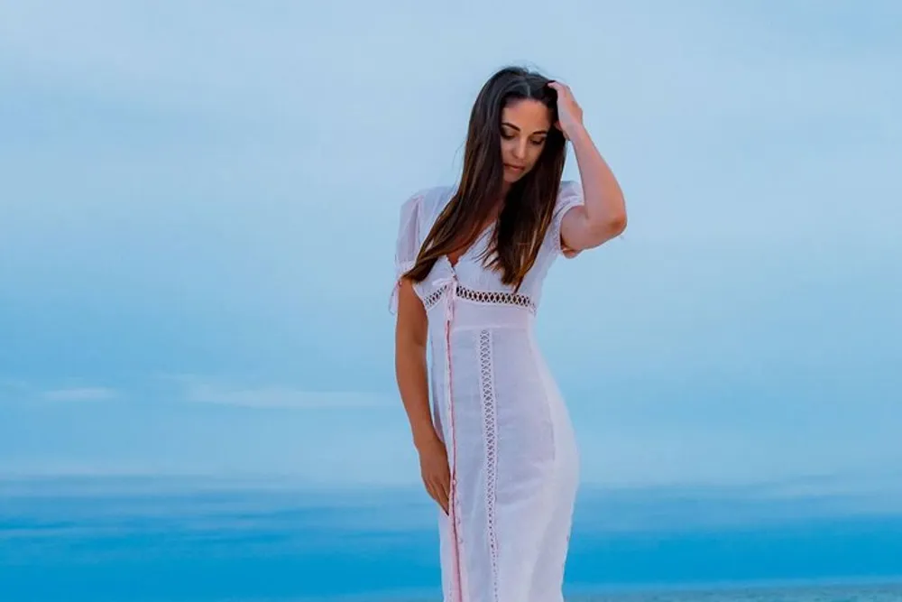 A woman in a white dress stands on a beach with a contemplative stance against a dusky blue sky