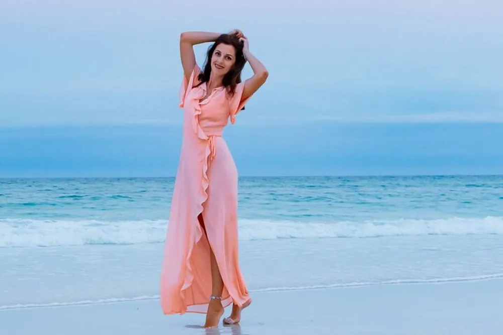A woman in an elegant peach dress is posing on the beach with one hand in her hair and a soft smile with the ocean in the background