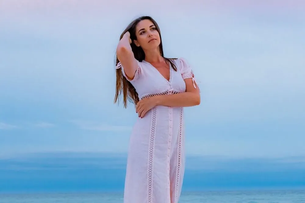 A woman in a white dress is posing on a beach with a pensive expression her hand in her hair against a soft blue sky