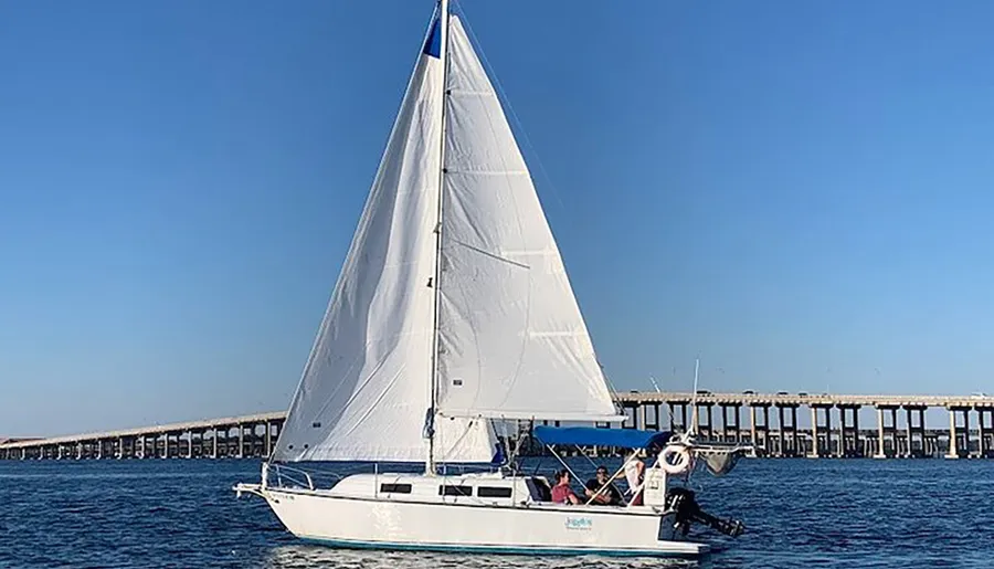 A sailboat with its sails unfurled glides across blue water near a long pier under a clear sky.
