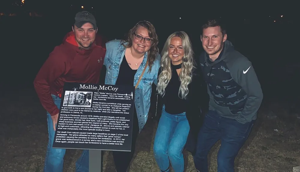 Four individuals are smiling for a photo at night with one of them holding a sign about a historical figure named Mollie McCoy