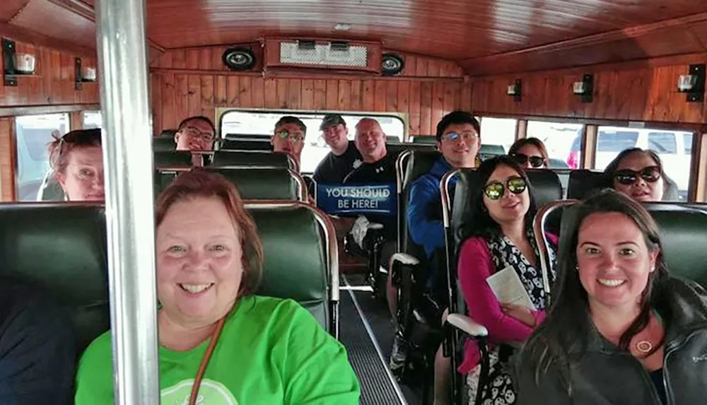 A group of smiling people are seated inside a vehicle with wooden interior posing for a photo with one person holding a sign that says YOU SHOULD BE HERE