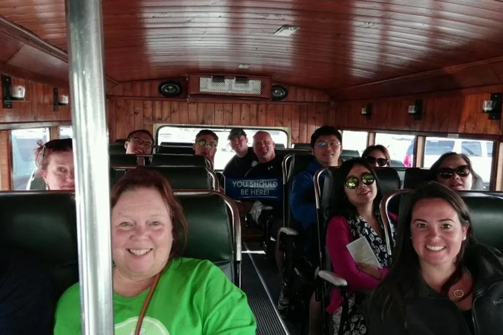 A group of smiling people is sitting inside a vintage-style bus with wooden interior paneling